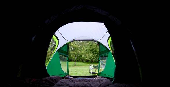 Blick von innen aus einem Zelt durch die offene Vorderseite auf einen Campingplatz mit Gras und Bäumen, mit einem Stuhl draußen.