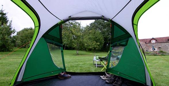 Drinnen in einem grünen Campingzelt, blickend durch die offene Tür auf einen Rasenplatz mit einem kleinen Tisch und Stuhl, Bäumen und einem Haus in der Ferne.