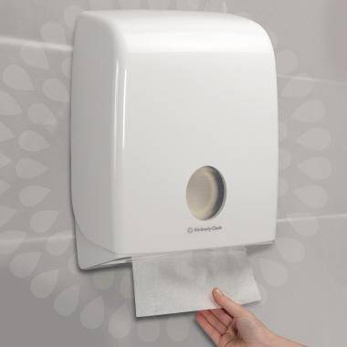 Ein an der Wand angebrachter Papierhandtuchspender mit einer Hand, die ein Handtuchblatt aus der unteren Öffnung heranzieht.