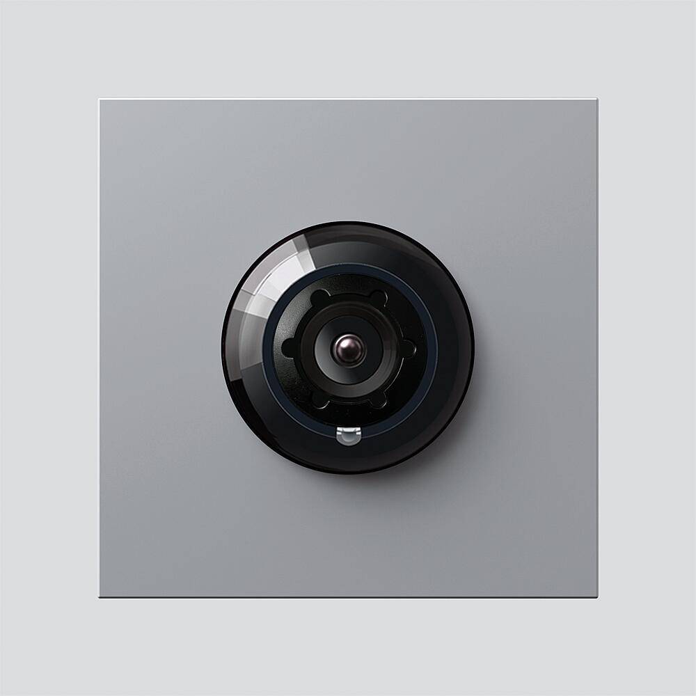 Eine kreisrunde schwarze Kameraobjektive, die in eine quadratische graue Oberfläche eingelassen ist. Die Linse reflektiert Licht und deutet darauf hin, dass sie möglicherweise eingeschaltet oder aktiv ist.