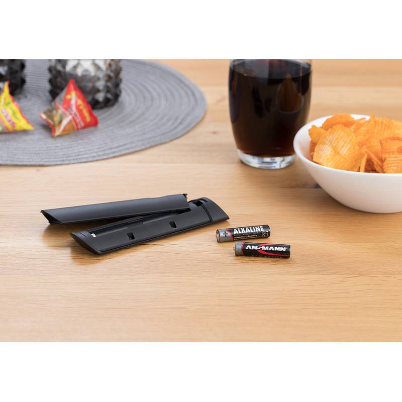Zwei AA-Batterien und ein schwarzer Batteriedeckel auf einem Tisch neben Chips, einem Getränk und Snacks auf einem Tischset.