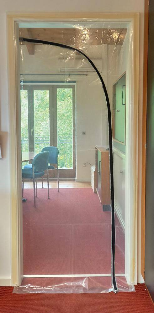 Türöffnung mit einem klaren Kunststoff-Streifenvorhang mit schwarzem gebogenem Rand, der einen Raum mit einem Stuhl und großem Fenster vom Flur trennt.