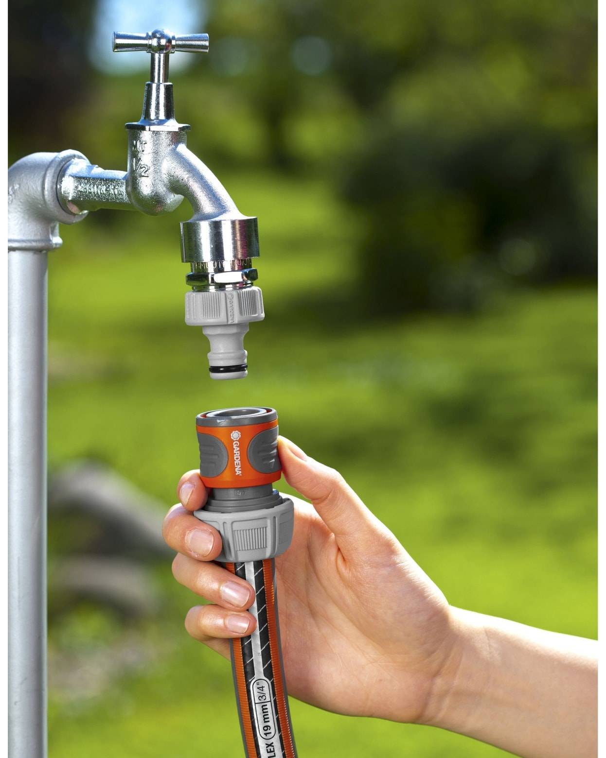 Hand, die eine Gartenschlauchdüse an einen Außenmetal-Wasserhahn anschließt, mit Wasserhahn oben und unscharfem grünem Rasen im Hintergrund.