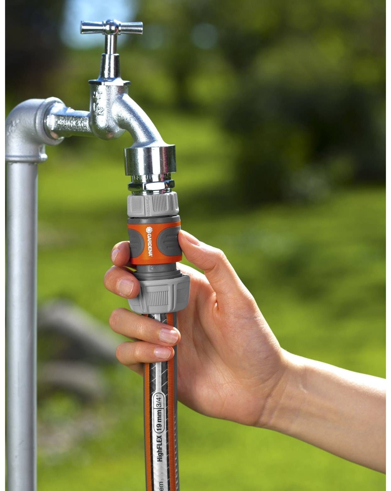 Hand, die einen Gartenschlauch mit einem Schlauchanschluss an einen Außenwasserhahn anschließt, mit unscharfem grünem Hintergrund.