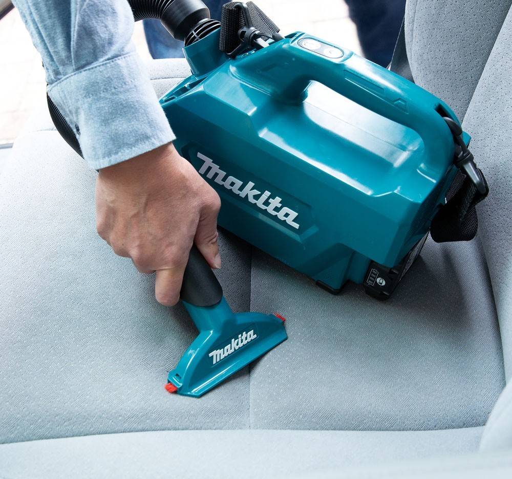 Eine Person verwendet einen blauen Staubsauger mit einem Markenschriftzug, um einen Autositz zu reinigen. Der Staubsauger ist kompakt und handlich, ideal für Polstermöbel.