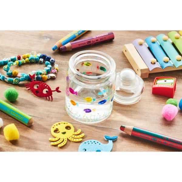 Bastelmaterialien auf einem Tisch: bemaltes Glasgefäß, farbige Perlen, Holzxylophon, Buntstifte, Pompons und kleine tierförmige Dekorationen.