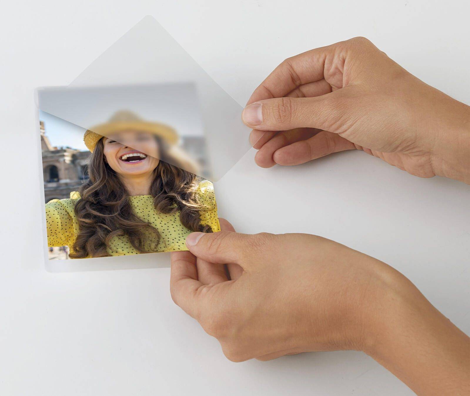 Hände schälen einen transparenten Schutzfilm von einem bedruckten Foto einer lächelnden Frau in einem Strohhut auf einer weißen Oberfläche ab.