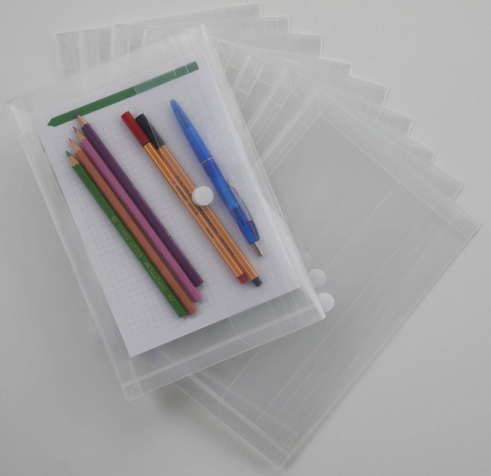 Ein Stapel durchsichtiger Kunststoff-Dokumentenhüllen mit einem Blatt Millimeterpapier und mehreren farbigen Bleistiften, Stiften und einem blauen Druckbleistift oben drauf.