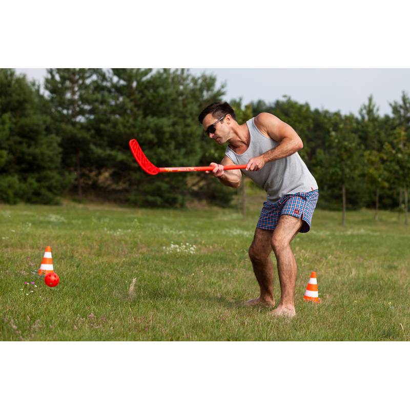 Eine Person in lockerer Kleidung und Sonnenbrille spielt Feldhockey auf Gras und verwendet einen roten Schläger in der Nähe von orangefarbenen Hütchen in einem Park.