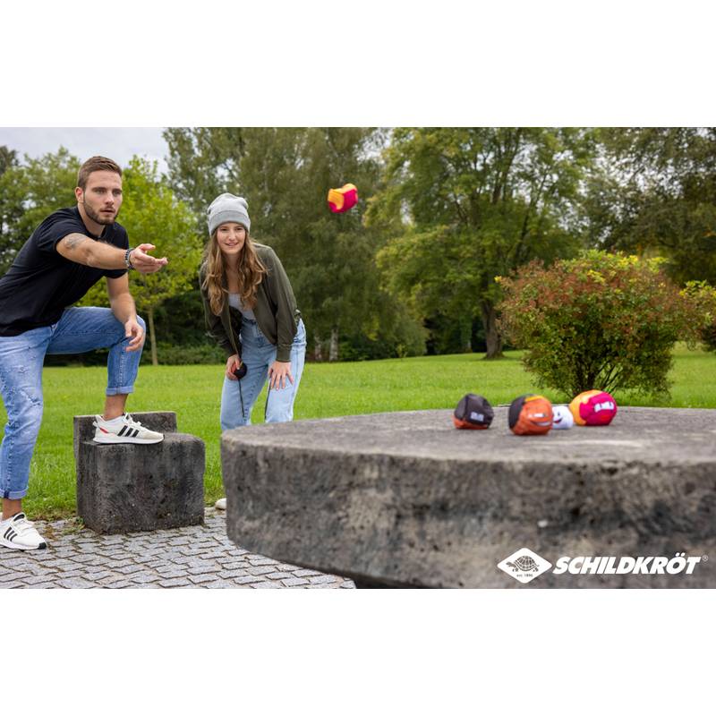 Ein Mann und eine Frau spielen Boccia mit bunten Kugeln auf einem steinernen Tisch in einer Parkanlage im Freien, mit Bäumen im Hintergrund.