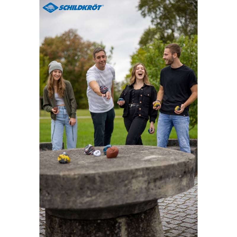 Vier Menschen werfen spielerisch Gegenstände auf einen runden Steintisch im Freien, lächeln und genießen eine Freizeitaktivität in einem Park.