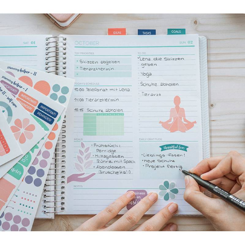 Offener Planer mit einer wöchentlichen Übersicht, auf der Aufgaben wie ‚Yoga' vermerkt sind, eine meditative Figur als Illustration und jemand, der ‚Projekt fertig' notiert.