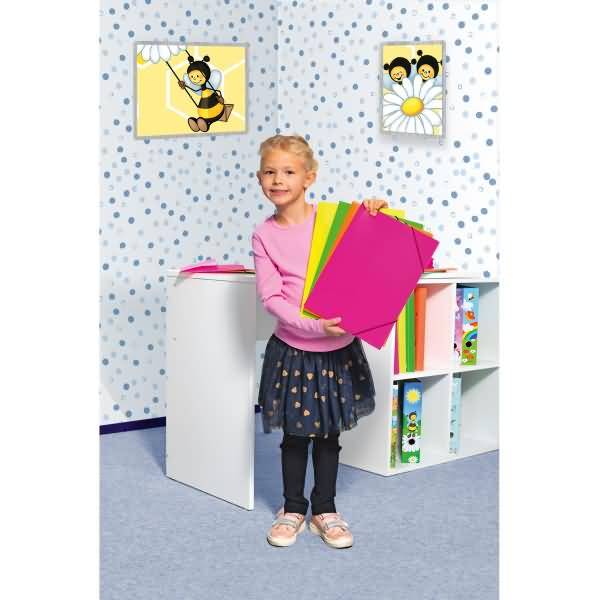 Junges Mädchen in einem Klassenzimmer, das einen Stapel farbenfroher Ordner hält, neben Fächern und Bienenmotiv-Wandkunst stehend.