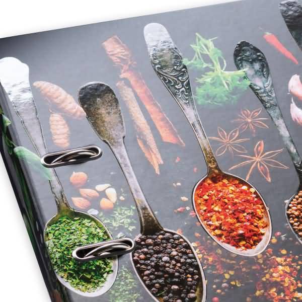 Nahaufnahme eines Spice-Themen-Ordners oder Notizbucheinbands mit übergroßen Löffeln gefüllt mit Kräutern und Gewürzen, gehalten durch Metallclips.