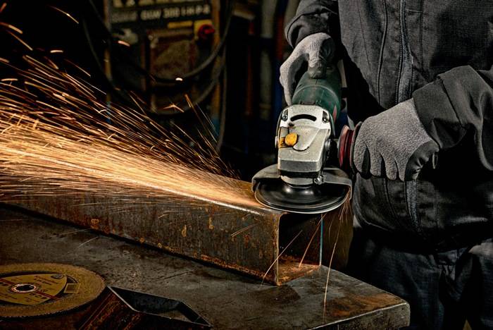 Person mit Handschuhen verwendet eine Winkelschleifer und erzeugt Funken, während er in einer Werkstattumgebung ein Metallstück zerschneidet.