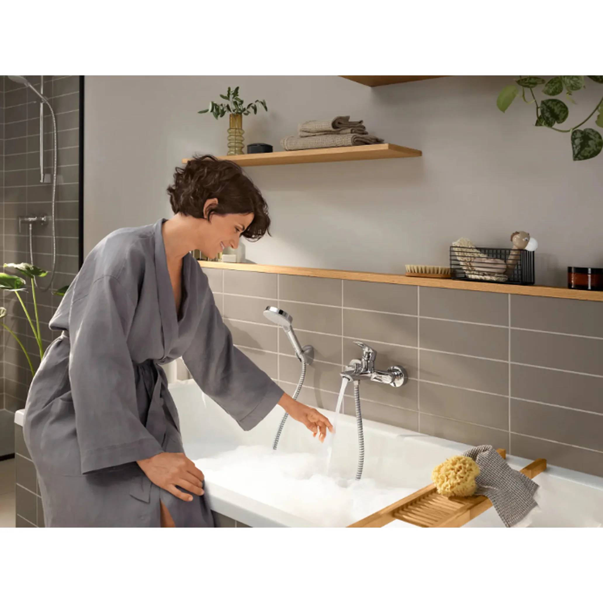 Eine Frau in einem grauen Bademantel beugt sich über eine mit Schaum gefüllte Badewanne und greift zum Wasser in einem modernen Badezimmer.