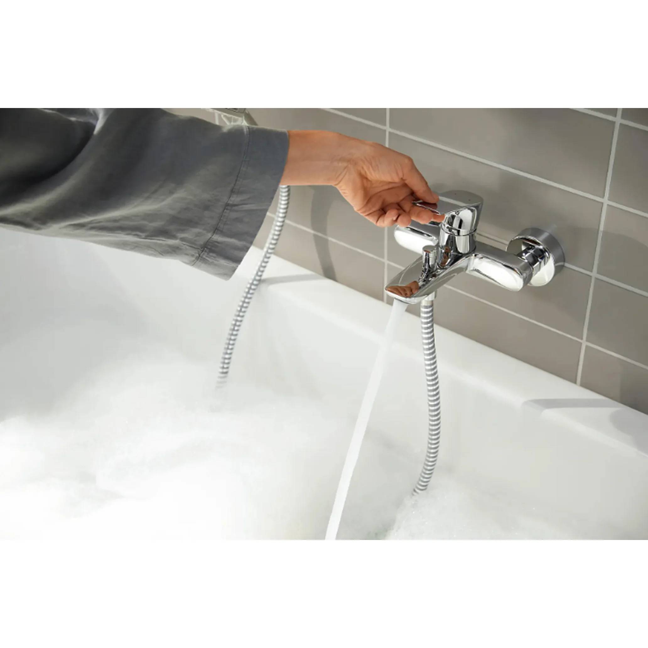 Hand dreht an einem chromfarbenen Badewannenhahn, fließendes Wasser füllt eine weiße Badewanne gegen eine Wand mit grauen Fliesen.