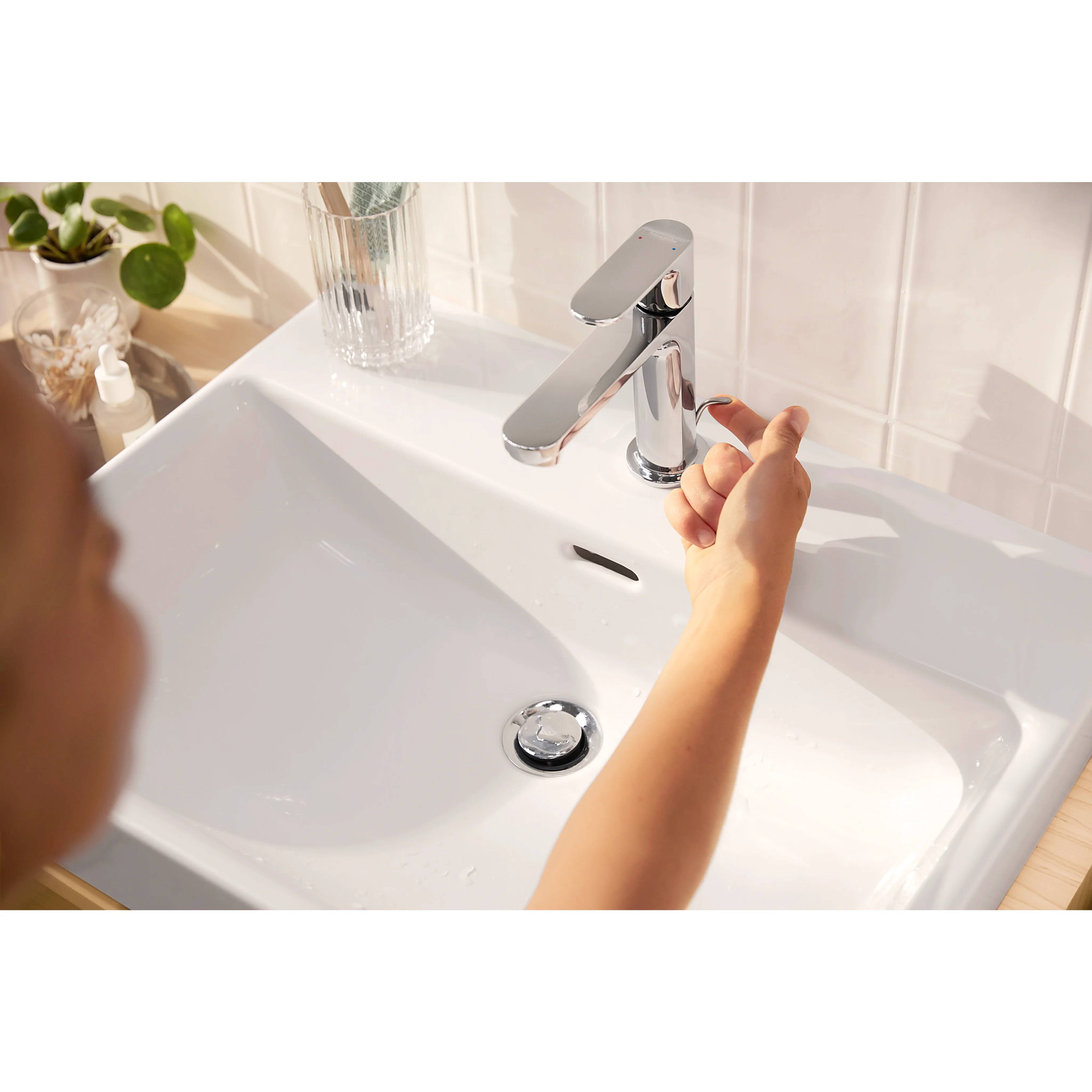 Kind schaltet einen verchromten Wasserhahn über einem weißen Waschbecken in einem hellen Badezimmer ein.