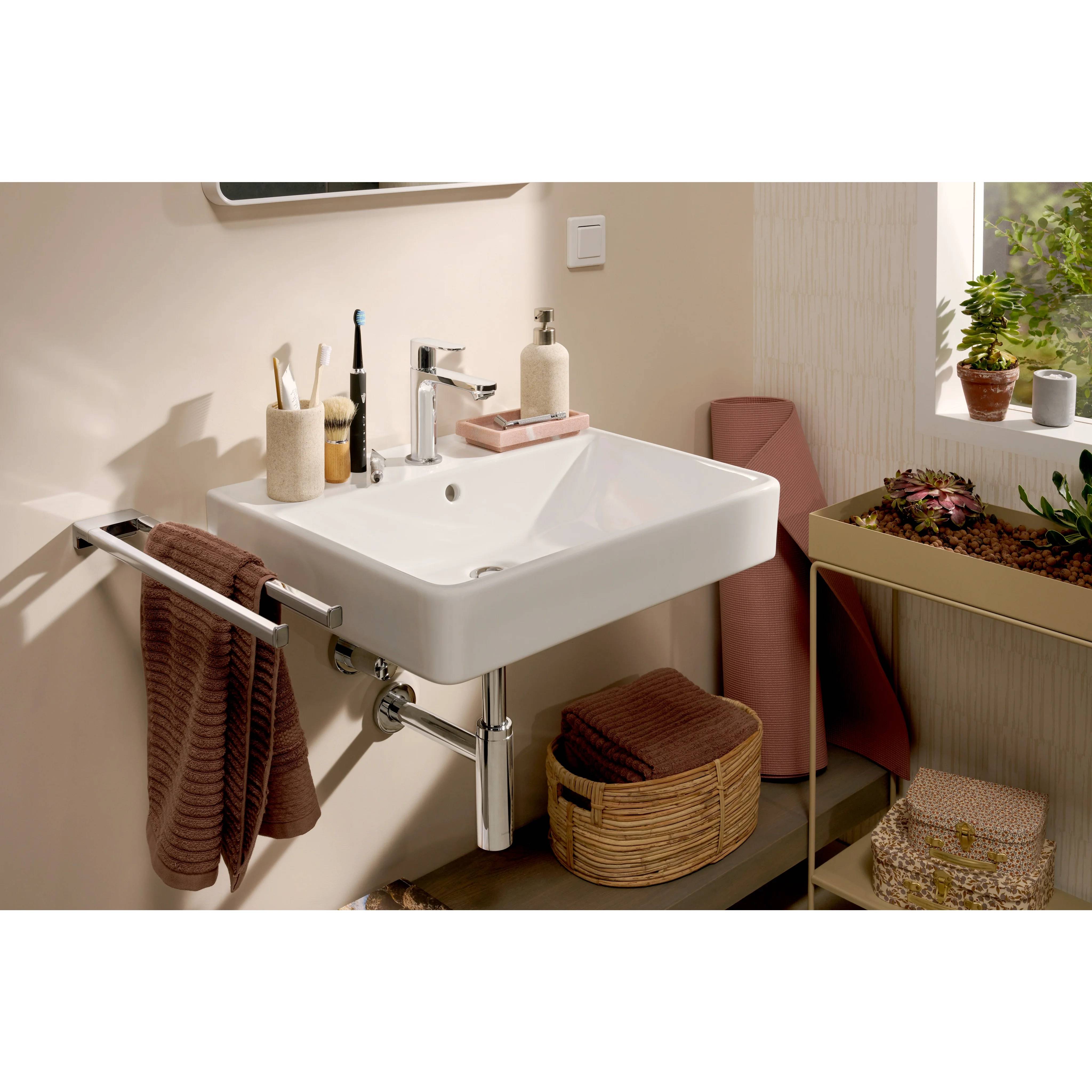 Ein kompaktes Badezimmerwaschtisch mit weißem Waschbecken, Handtuchhalter, Toilettenartikeln, Aufbewahrungskorb und Topfpflanzen neben einem Fenster.