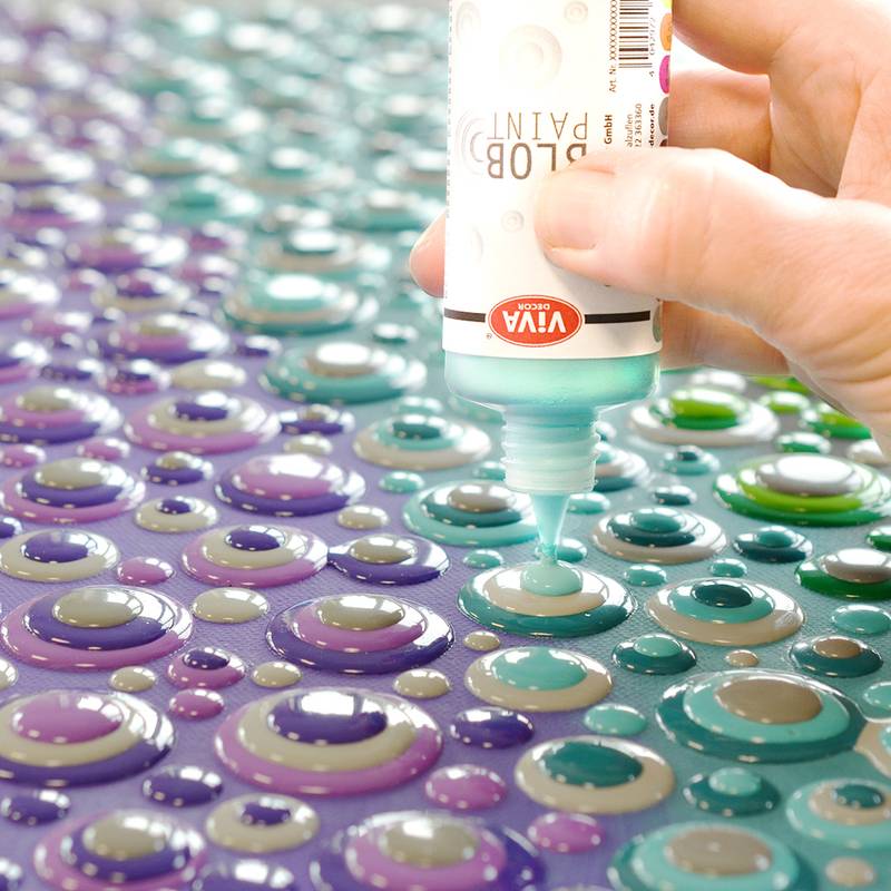 Eine Hand verwendet eine Tube mit farbiger Farbe, um 3D-Tropfen auf einer violetten Oberfläche zu erzeugen und dabei ein strukturiertes, dekoratives Muster in verschiedenen Nuancen zu bilden.