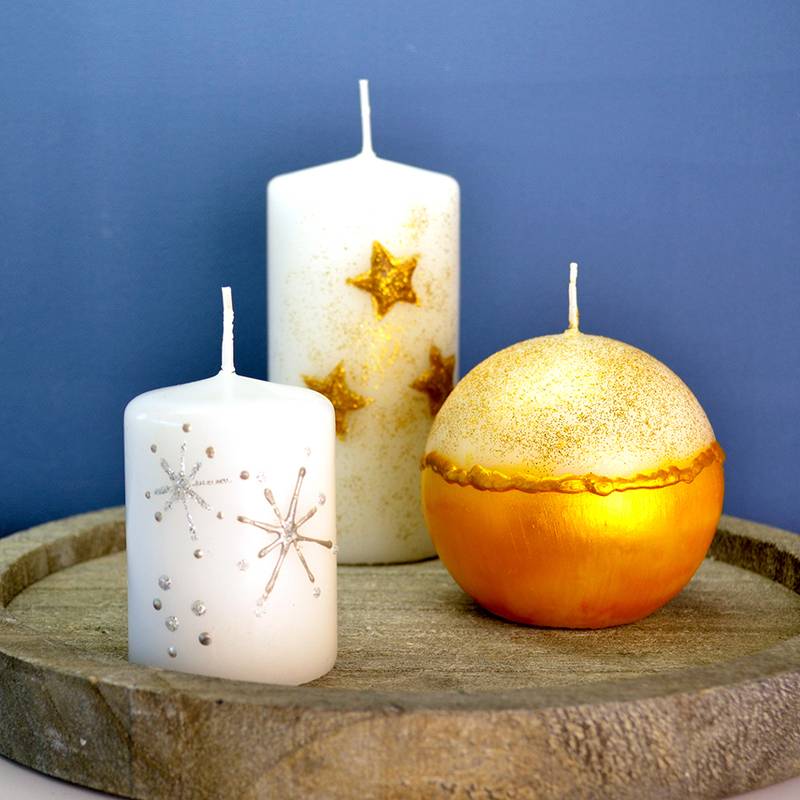 Drei dekorative Kerzen auf einem Holztablett: eine weiße Kerze mit silbernen Sternen, eine weiße Kerze mit goldenen Sternen und eine runde goldene Kerze.