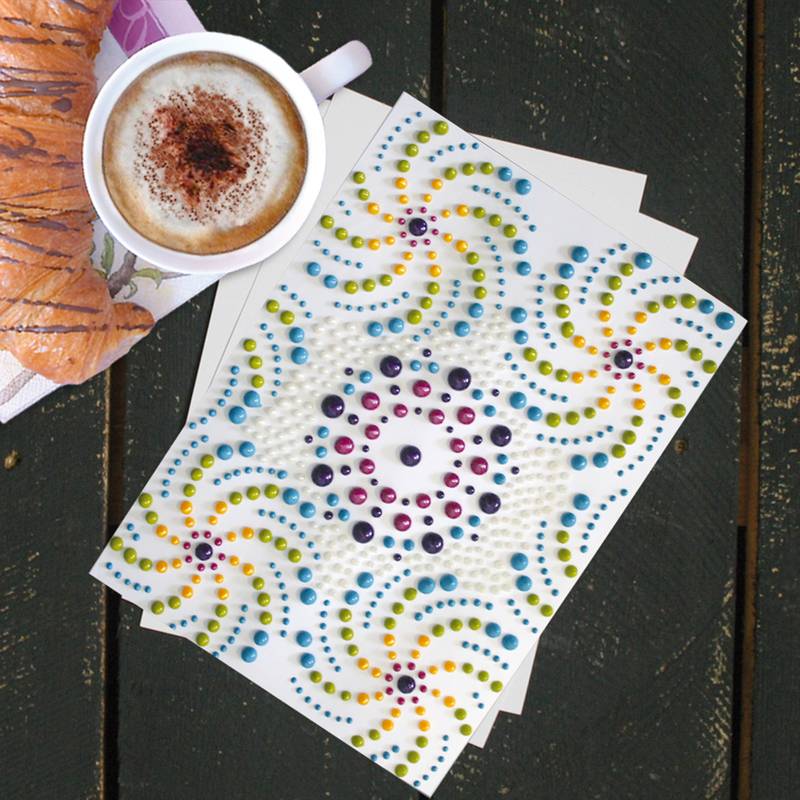 Ein verziertes Papier mit bunten Punktmustern liegt auf einem Tisch neben einem Cappuccino mit schaumiger Milch und Schokoladenstreusel sowie einem Croissant.