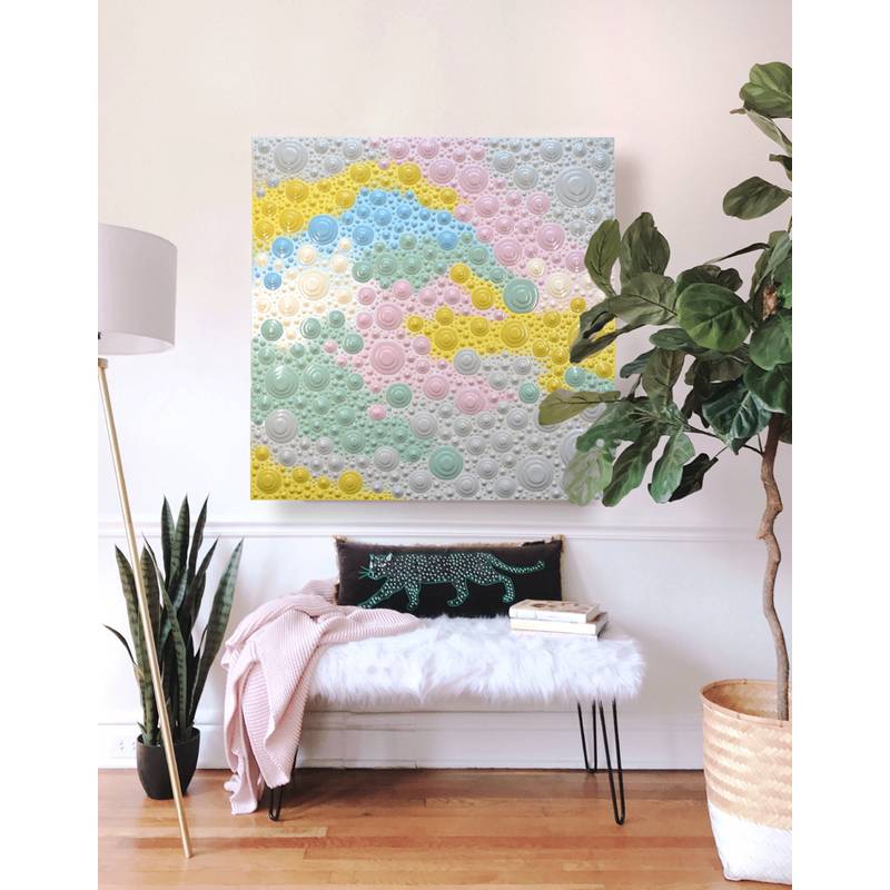 Ein gemütlicher Raum zeigt eine weiße, pelzige Bank mit einer rosa Decke und gemusterten Kissen. Darüber hängt an der Wand ein farbenfrohes abstraktes Kunstwerk.