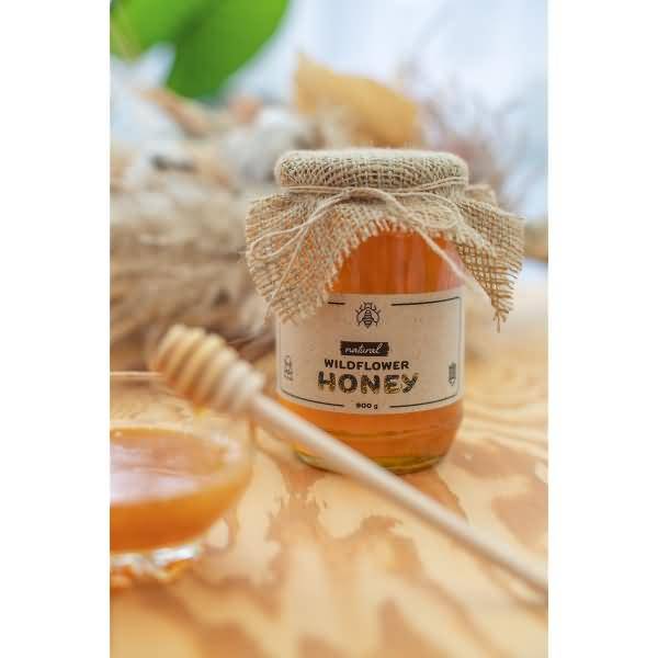 Glas mit Wildblütenhonig mit Juteband-Verpackung und Etikett, gezeigt auf einem Holztisch mit einem Honigschöpfer und einer Schüssel mit Honig.