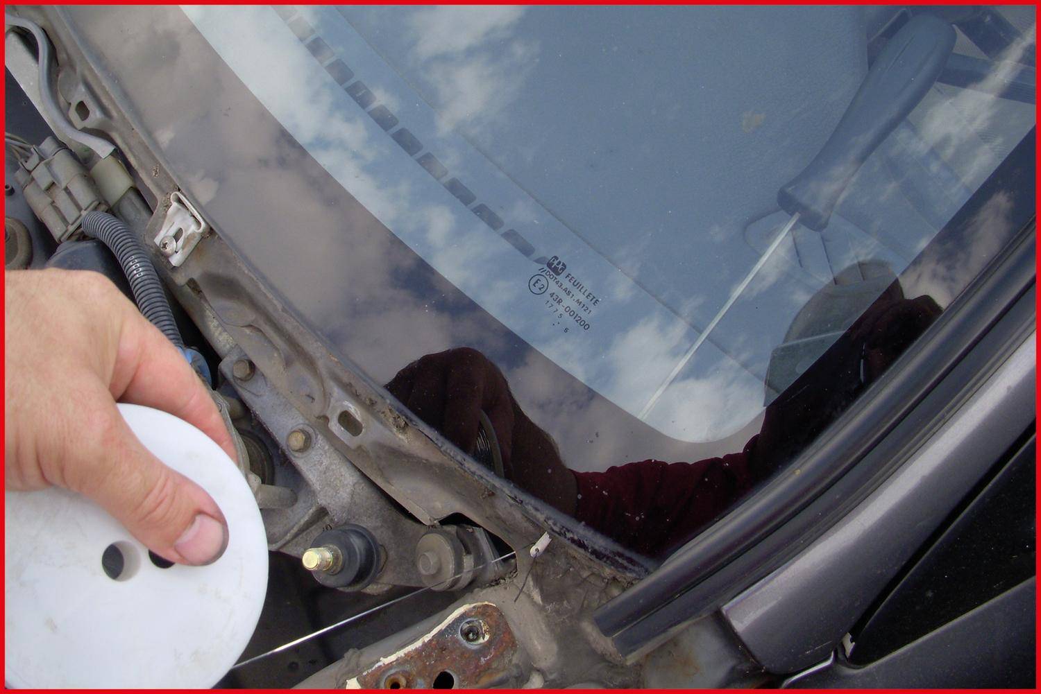 Eine Person, die Flüssigkeit in einen Scheibenwaschflüssigkeitsbehälter eines Autos gießt, mit offener Motorhaube und teilweise sichtbarem Windschutzscheibenbereich.