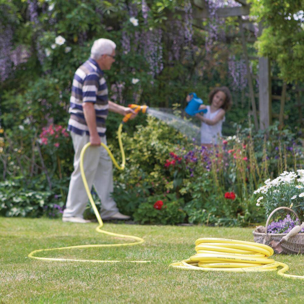 Ein älterer Erwachsener und ein Kind gießen Gartenpflanzen mit einem Gartenschlauch an einem sonnigen Tag, umgeben von leuchtenden Blumen und üppigem Grün.