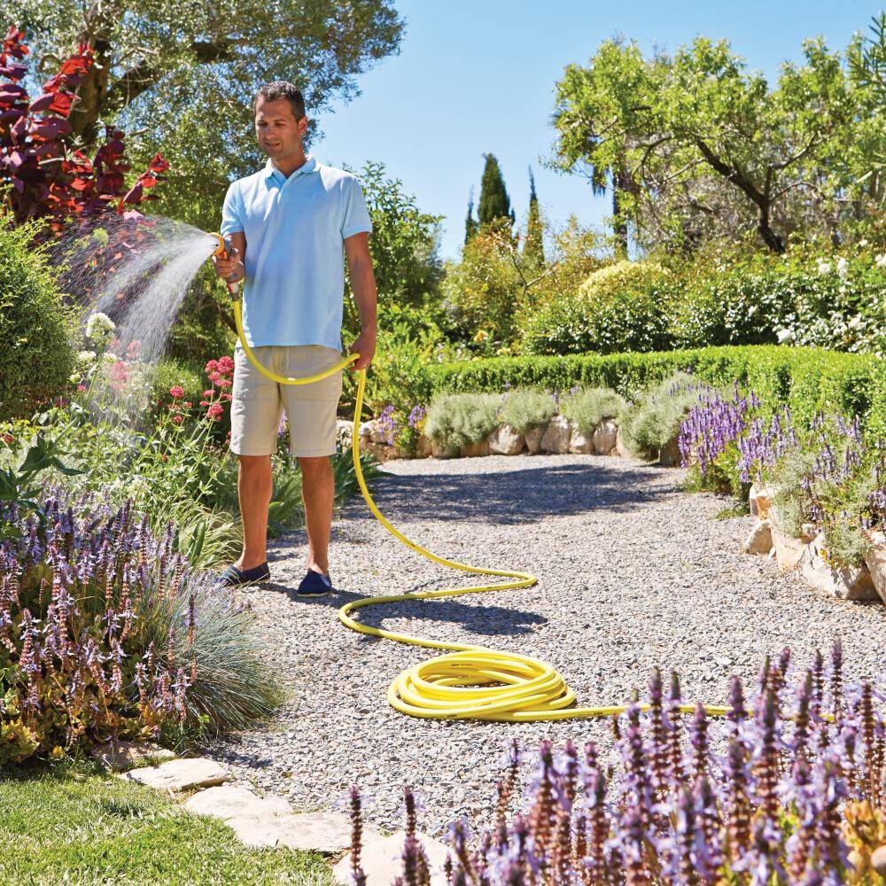 Eine Person, die an einem sonnigen Tag einen Garten mit einem Gartenschlauch bewässert, umgeben von verschiedenen Pflanzen und Blumen. Ein Kiesweg schlängelt sich durch den Garten.
