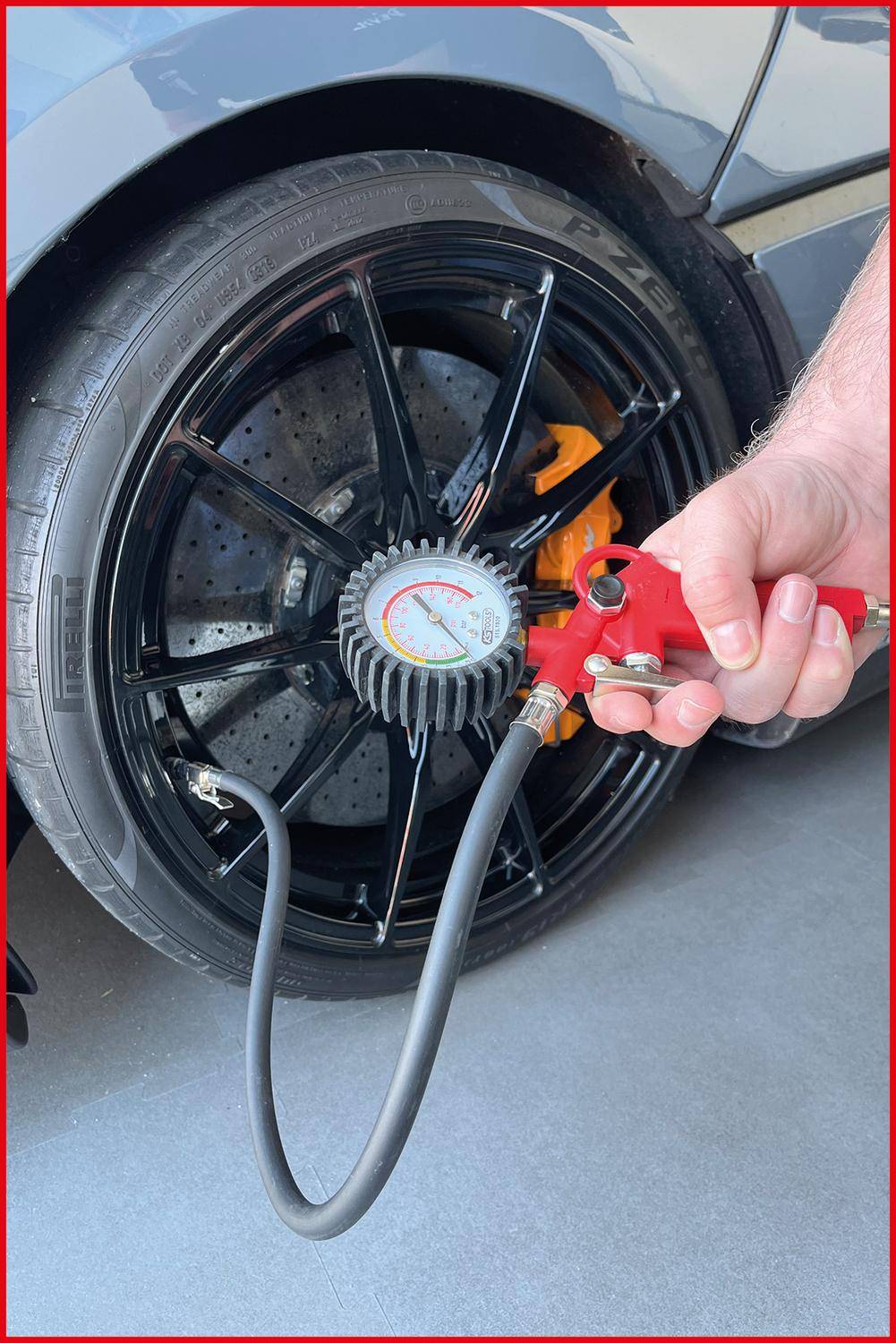 Eine Person prüft den Luftdruck eines Autoreifen mit einem Manometer, das an einer roten Luftpumpe befestigt ist. Der Reifen weist ein sichtbares Profil auf.