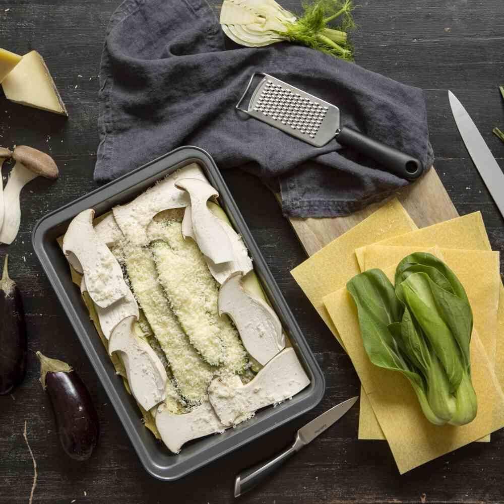 Eine Auflaufform mit geschichteten Auberginen und Lasagneblättern, die mit Ricotta-Käse bedeckt ist, umgeben von frischen Zutaten und Küchenutensilien.