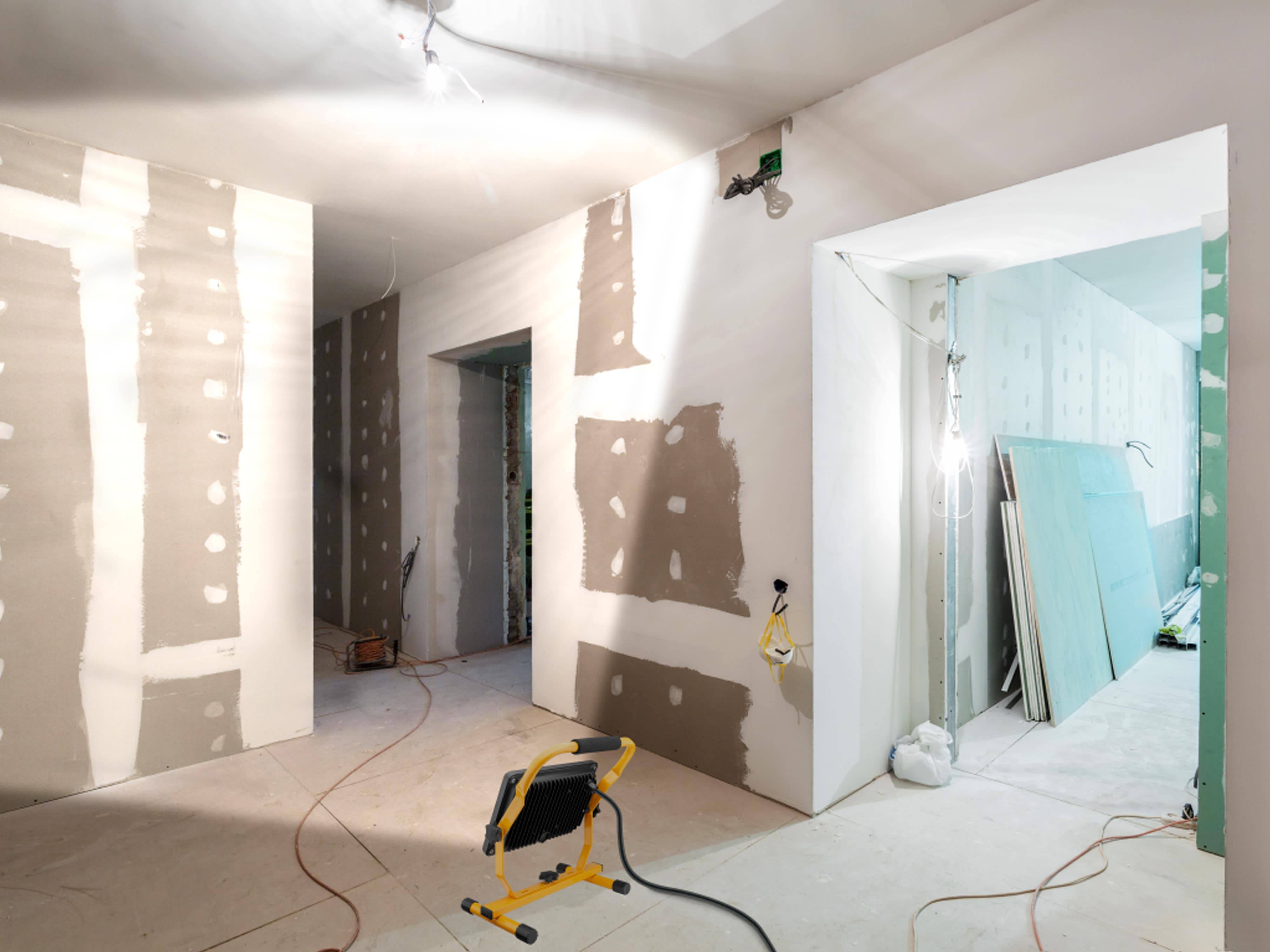 Ein Renovierungsraum mit teilweise mit Spachtelmasse verkleideten Wänden, freigelegten Leuchten, einer Arbeitsleuchte auf dem Boden und Baumaterialien.