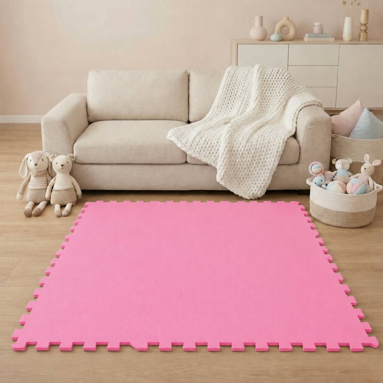 Wohnzimmer mit einem beigen Sofa, einer Strickdecke, Spielzeugen und einer leuchtend rosa Schaumstoff-Spielmatte in der Mitte des Bodens.