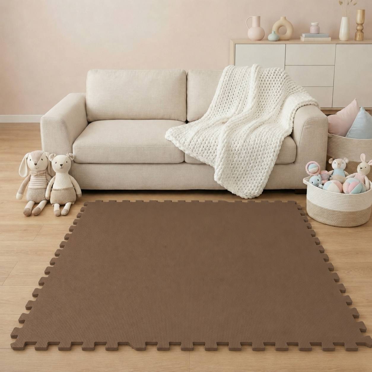 Ein gemütliches Wohnzimmer mit einem beigefarbenen Sofa, einer weißen Strickdecke, Plüschtieren und einer großen braunen Schaumstoff-Spielmatte auf dem Boden.