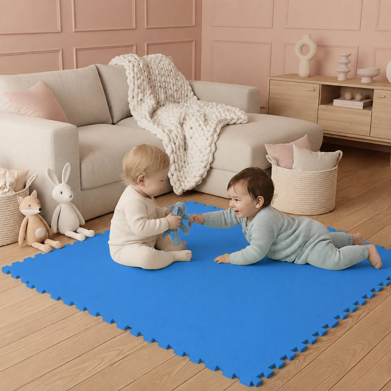 Zwei Kleinkinder spielen sich gegenseitig zugewandt auf einer blauen Schaumstoffmatte in einem gemütlichen Wohnzimmer mit einem cremefarbenen Sofa, Kuscheltieren und einem Spielzeugkorb.