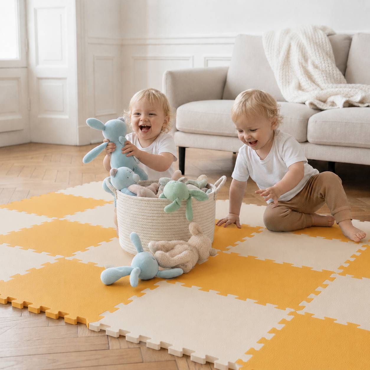 Zwei Kleinkinder spielen auf einer gelb-weißen Schaumstoffmatte und sortieren Kuscheltiere aus einem Korb in einem hellen Wohnzimmer.