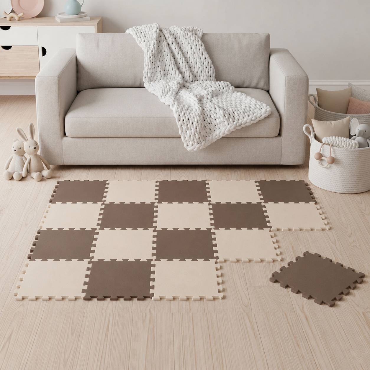Ein beiges Sofa mit einer gestrickten Wurfdecke steht hinter einer karierten Schaumstoff-Spielmatte auf einem hellem Holzboden, mit Spielzeugen in Körben in der Nähe.