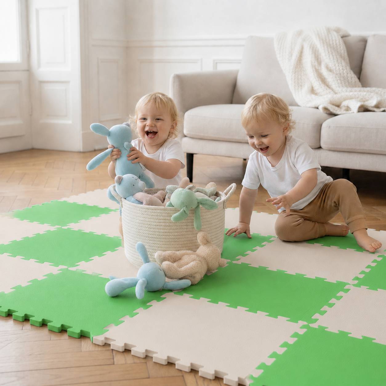 Zwei Kleinkinder spielen auf einer Schaumstoffmatte, lächeln und handhaben Kuscheltiere neben einem Korb in einem hellen Wohnzimmer.