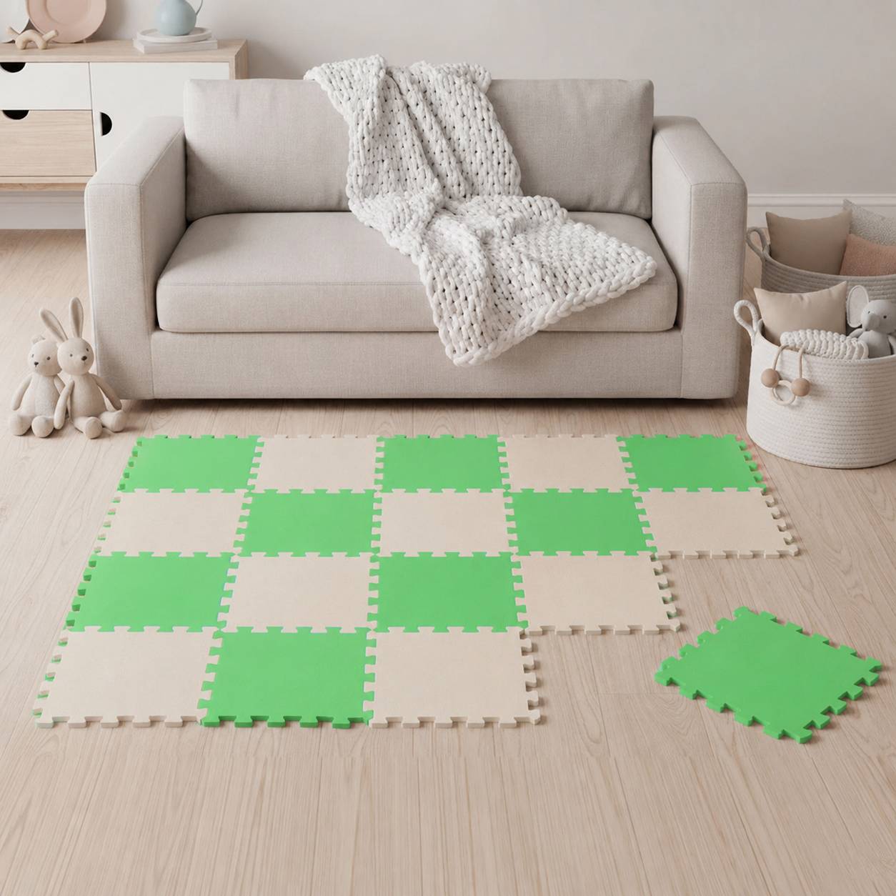 Gemütliches Wohnzimmer mit einem beigen Sofa und gestrickter Wurfdecke; grüne und cremefarbene Schaumstoff-Spielmatten bedecken den Boden, mit Spielzeugen und Aufbewahrungskörben in der Nähe.