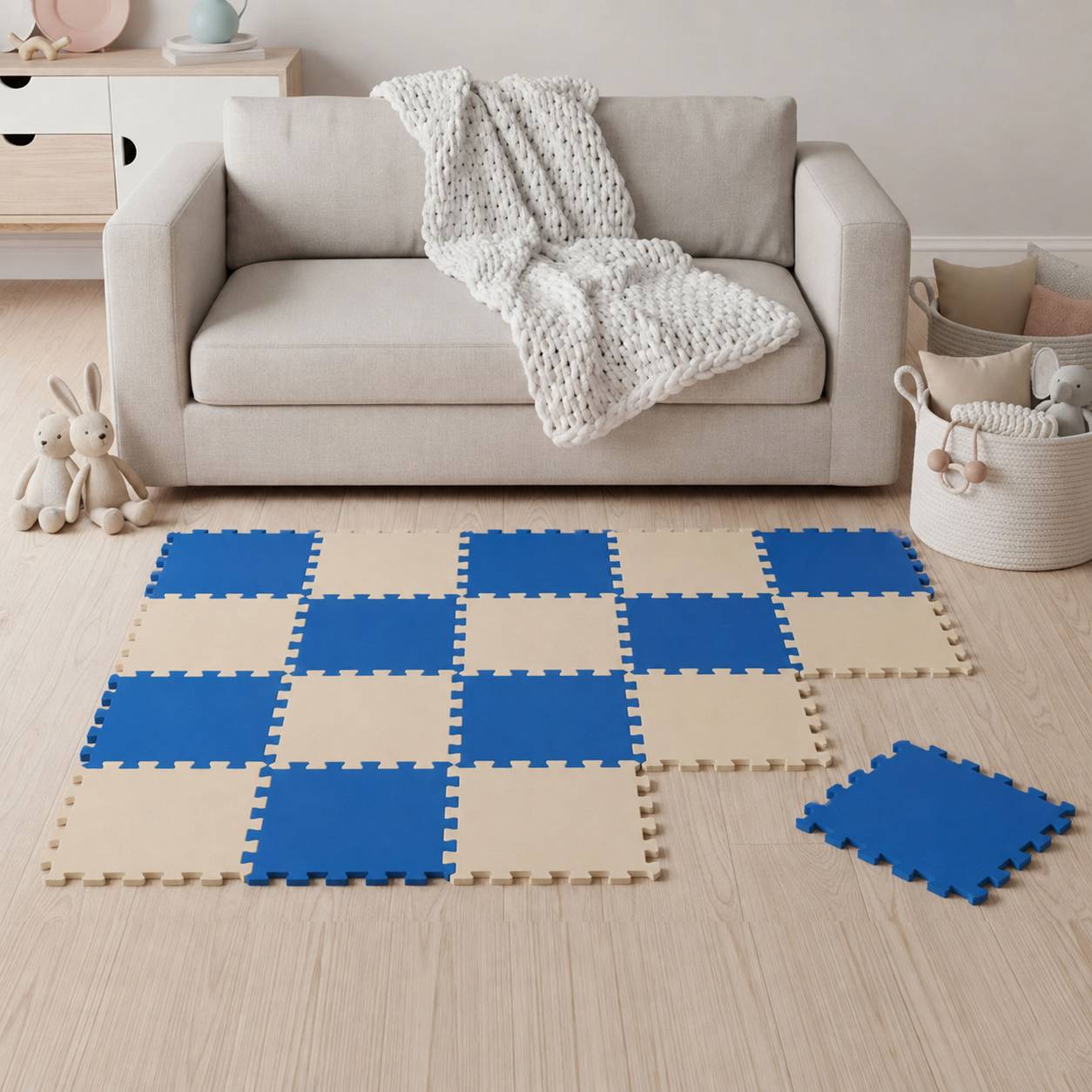 Ein beiges Sofa mit einer gestrickten Decke sitzt hinter blau-beige Schaumstoff-Spielmatten, die auf dem Boden angeordnet sind, mit Spielzeugen und einem Aufbewahrungskorb in der Nähe.