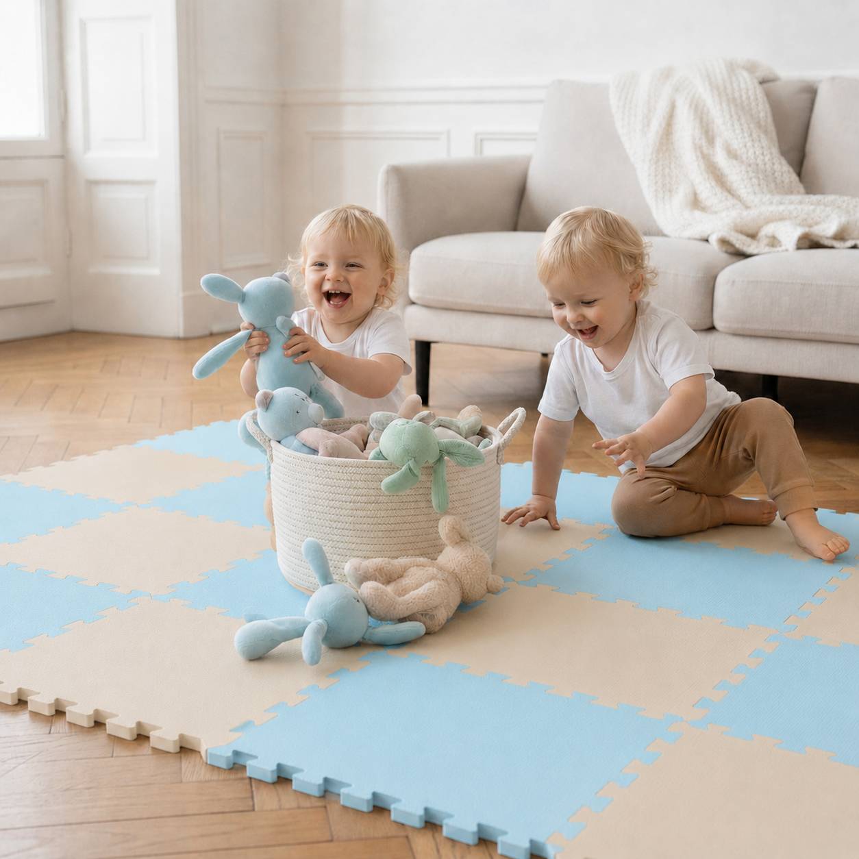 Zwei Kleinkindern spielen mit Kuscheltieren auf einer Schaumstoffmatte in einem hellen Wohnzimmer, neben einem Spielzeugkorb und einem Sofa.