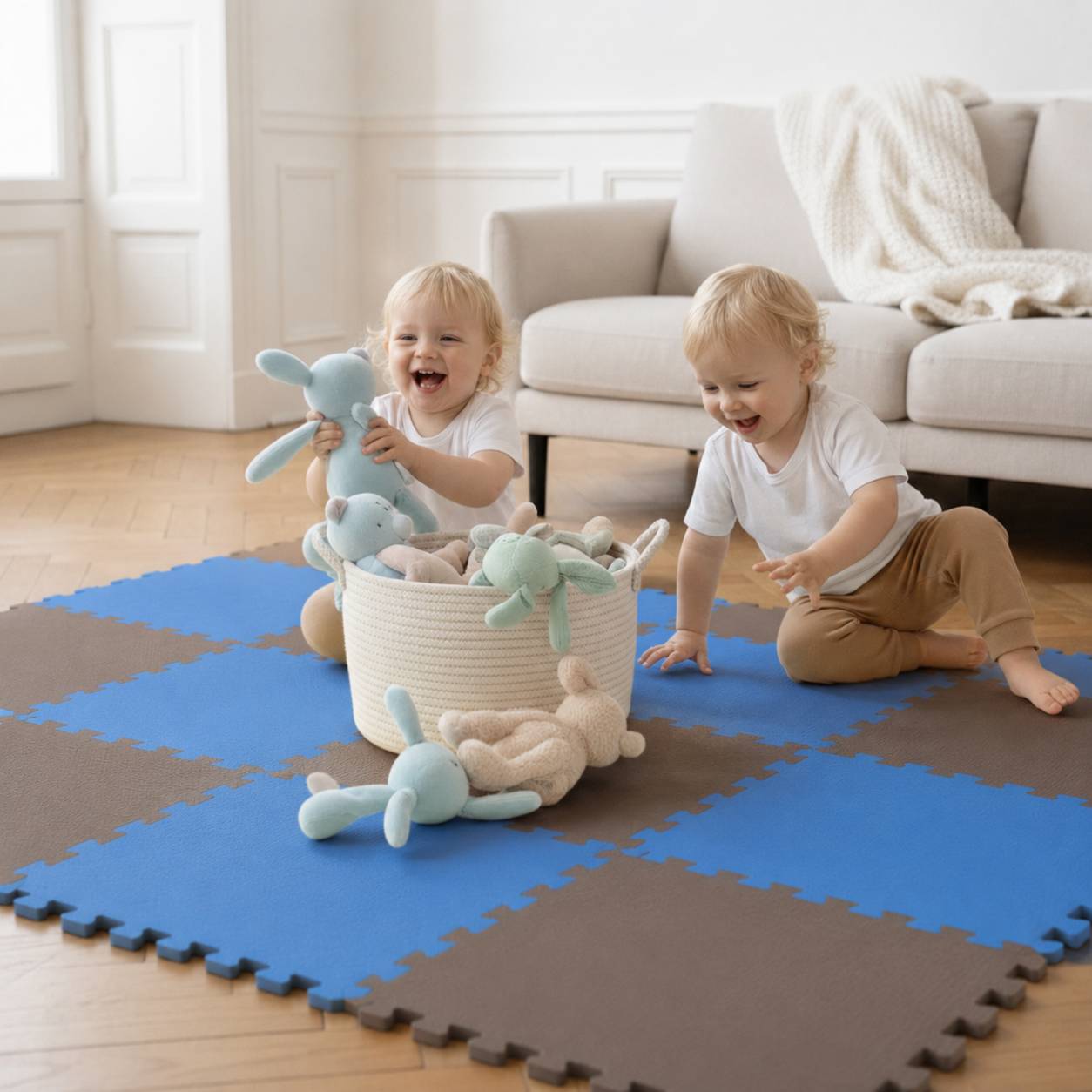 Zwei Kleinkinder spielen auf einer Schaumstoffmatte, lächeln und greifen nach Plüschhäschen neben einem Spielzeugkorb in einem hellen Wohnzimmer.