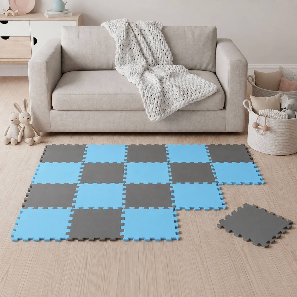 Wohnzimmer mit einem beigen Sofa und gestrickter Decke, mit verschachtelten blauen und grauen Schaumstoff-Bodenfliesen, die wie eine karierte Spielmatte angeordnet sind, mit einer losen Fliese in der Nähe.