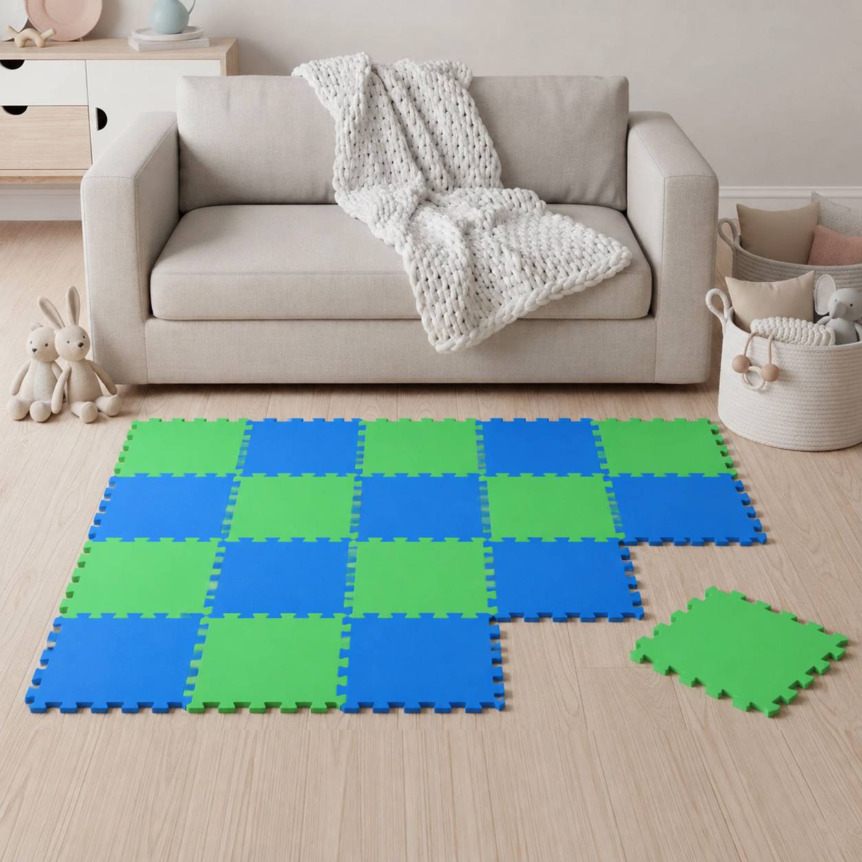 Wohnzimmer mit einem beigefarbenen Sofa und einer gestrickten Wurfdecke, auf dem Boden sind verschlungene blaue und grüne Schaumstoff-Bodenfliesen verlegt, daneben liegt eine lose Fliese.