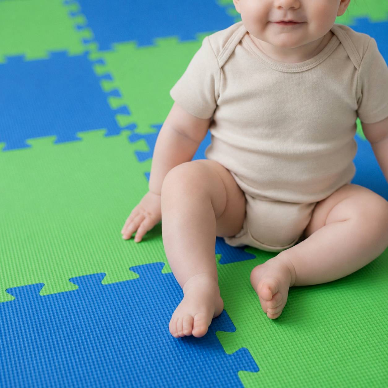 Baby sitzt auf grünen und blauen Schaumstoff-Spielmatten und trägt einen beigefarbenen Strampler.