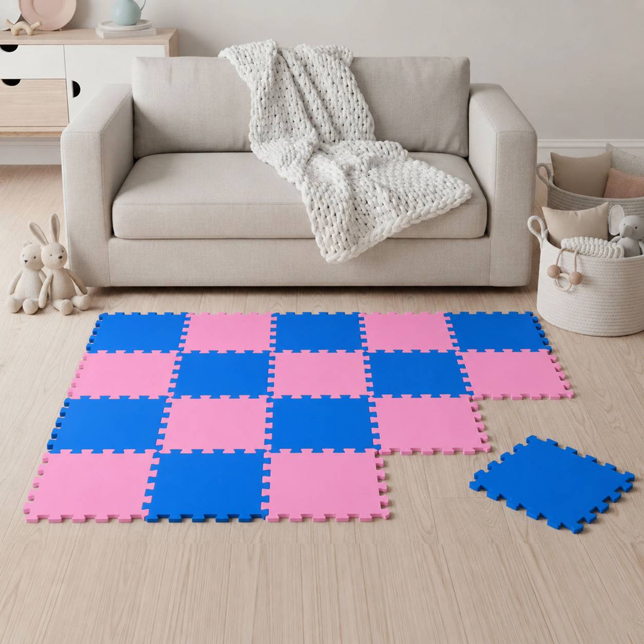 Rosa und blaue ineinander greifende Schaumstoff-Bodenfliesen, angeordnet in einem Spielbereich vor einem beigen Sofa mit einer weißen Wurfdecke.