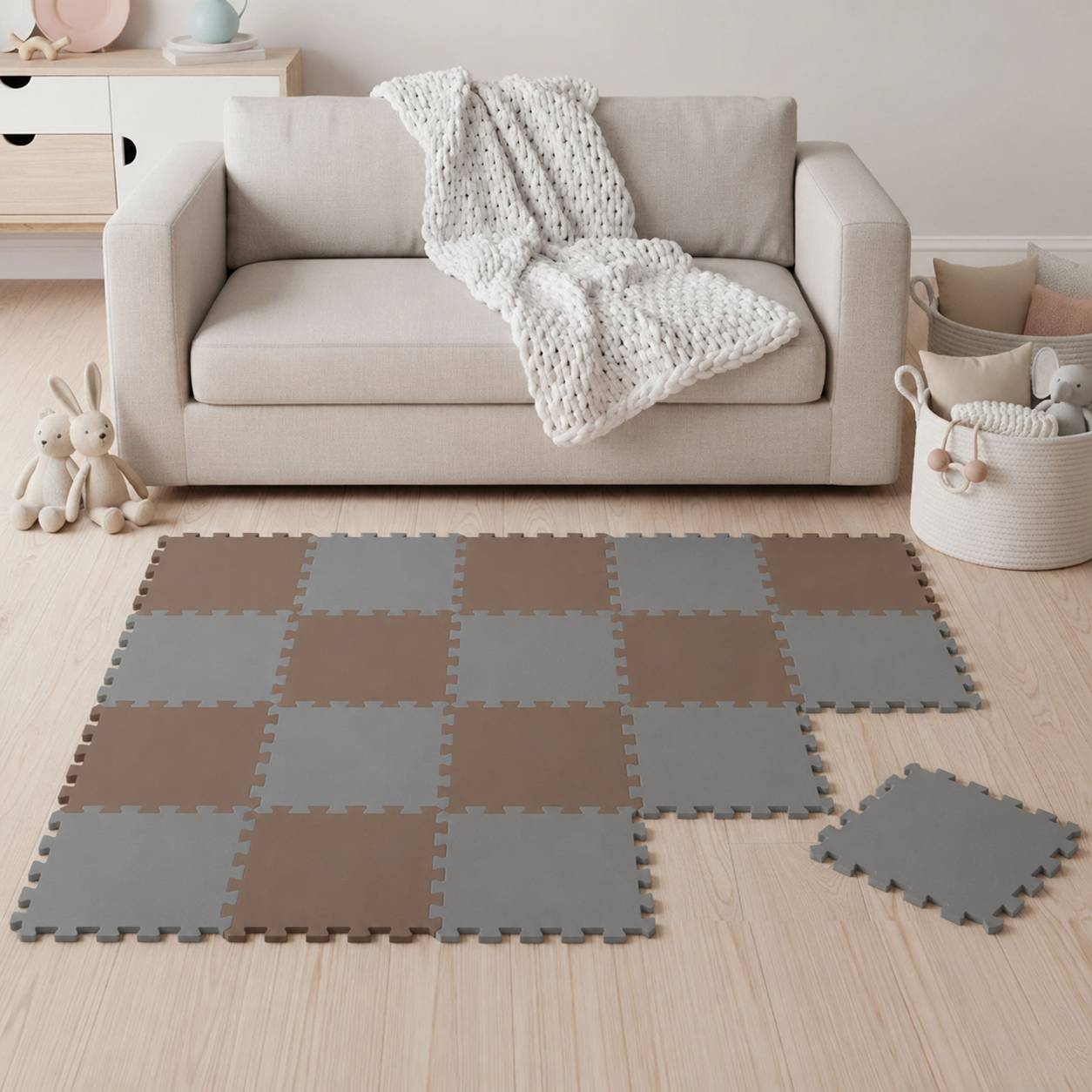 Wohnzimmer mit einem beigen Sofa, einer Strickdecke, Spielzeugen und Aufbewahrungskörben sowie einer graubraun gemusterten verschlungenen Schaumstoff-Spielmatte auf dem Boden.
