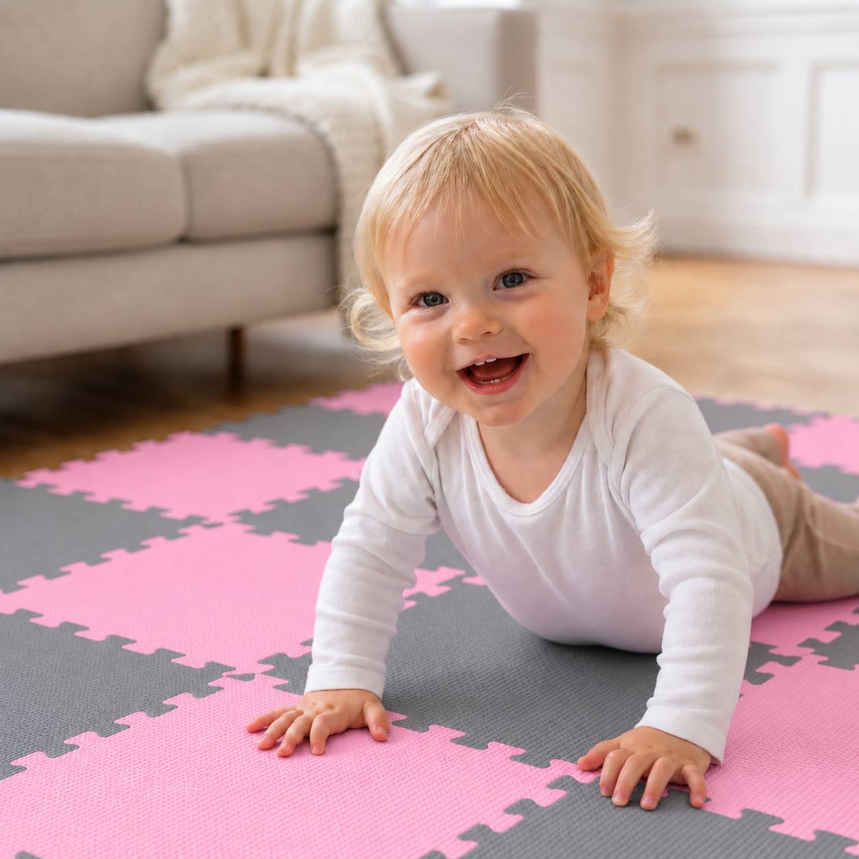 Ein lächelndes blondes Baby krabbelt auf einer rosa und grauen Schaumstoff-Spielmatte in einem hellen Wohnzimmer.